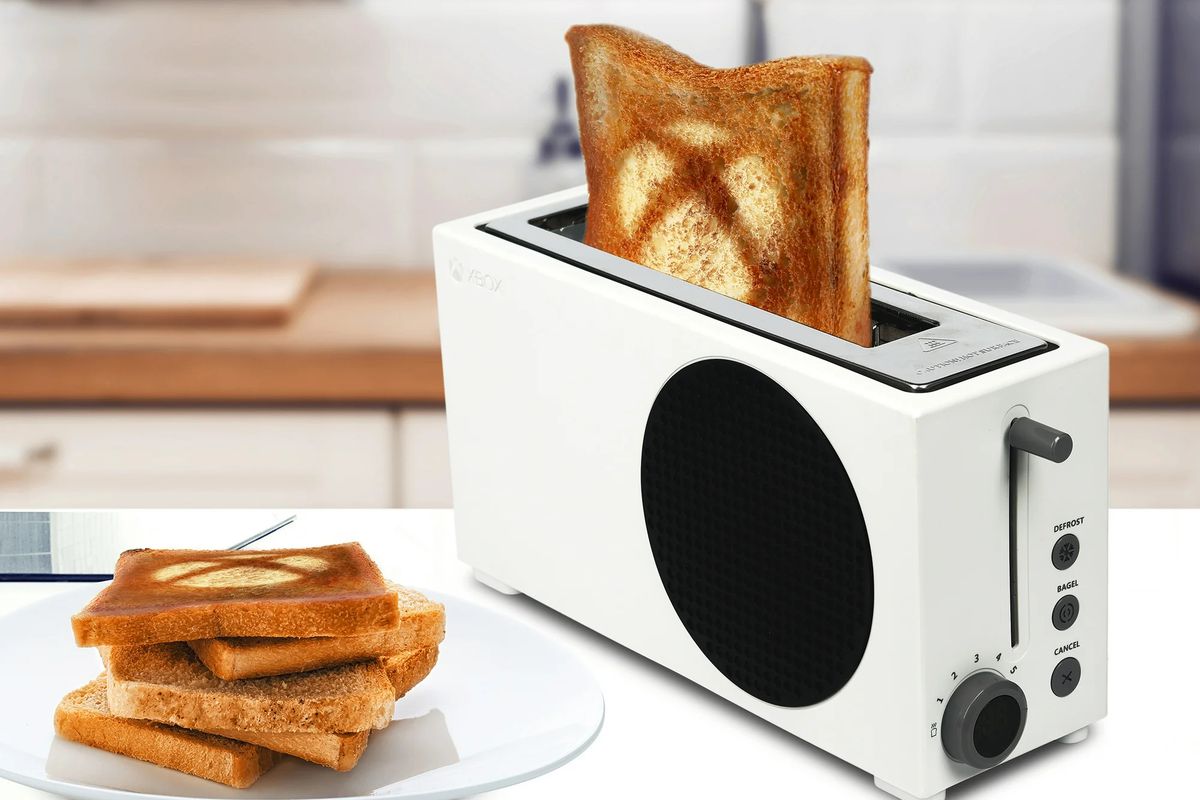 A photo of the Xbox Series S toaster next to a plate with toast stacked on it. Popping out of the toaster is a piece of prepared toast that has the Xbox logo printed on it from the toasters heating coils.