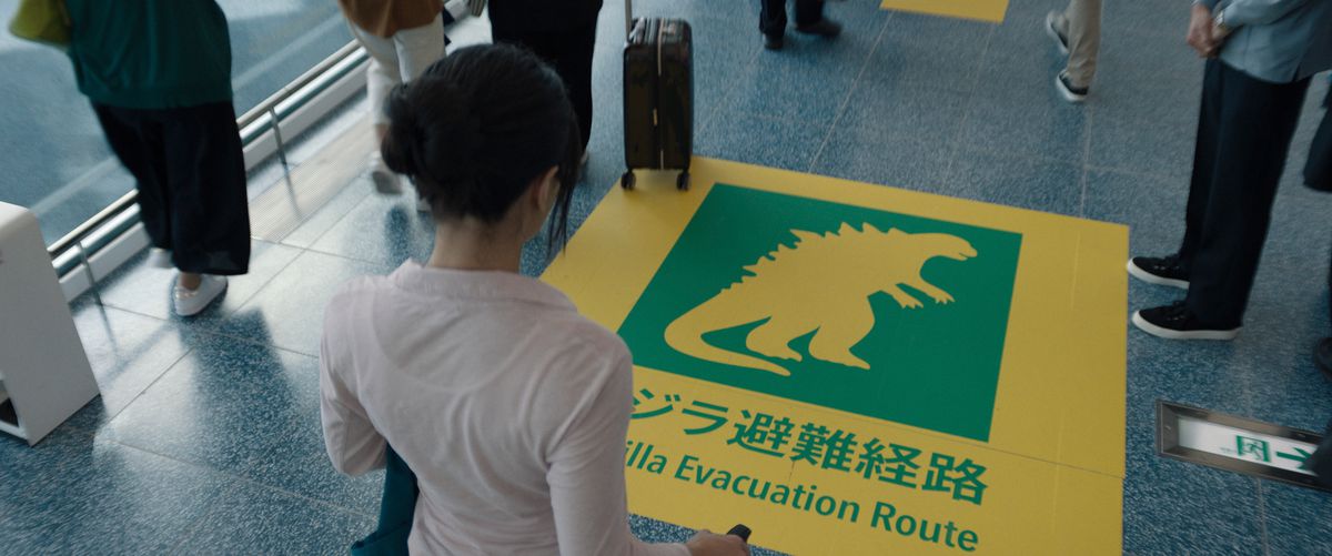 A woman stands in an airport in front of a sign on the floor marking a Godzilla evacuation route in the Apple TV Plus show Monarch: Legacy of Monsters