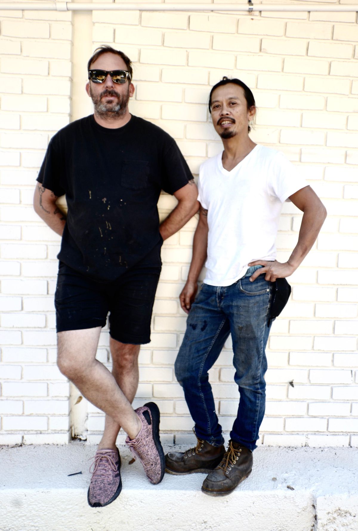 Skip Englebrecht in black tee and black shorts leans against a white brick wall next to chef Nhan Le in a white v-neck tee, jeans, and work boots