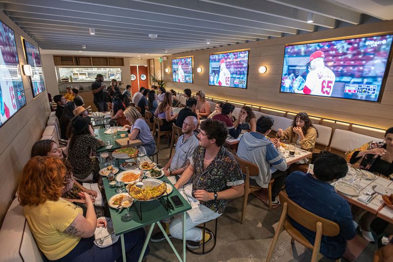 A dining room is full of young diners seated on banquettes and at small tables, with TV screens showing sports in the background. 