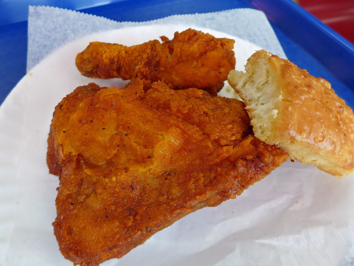 Two pieces of fried chicken with a biscuit on a white paper plate on a blue tray