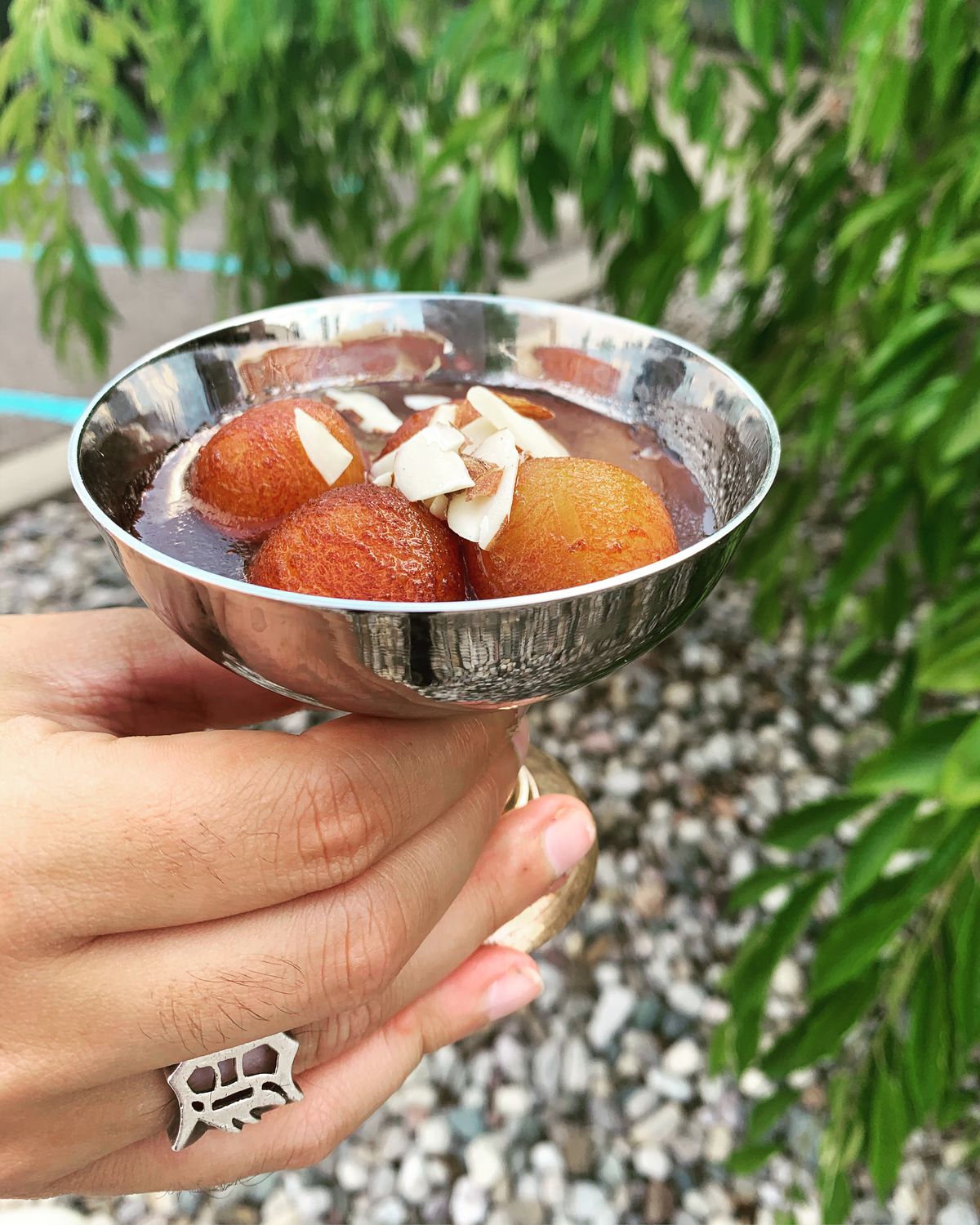 A hand holds a metal saucer filled with four gulab jamun covered with almond slivers.