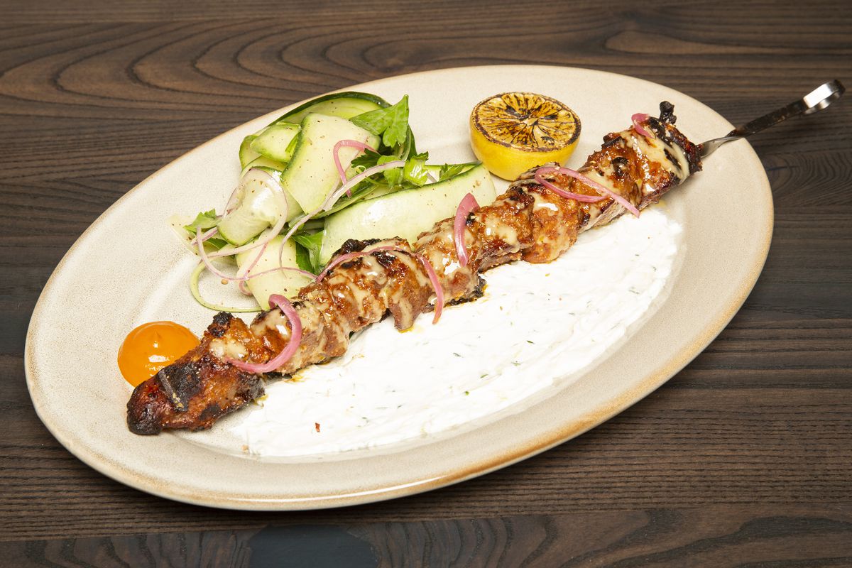 A long skewer of grilled chicken on an oval-shaped white plate. A small cucumber salad and grilled lemon sit beside it.