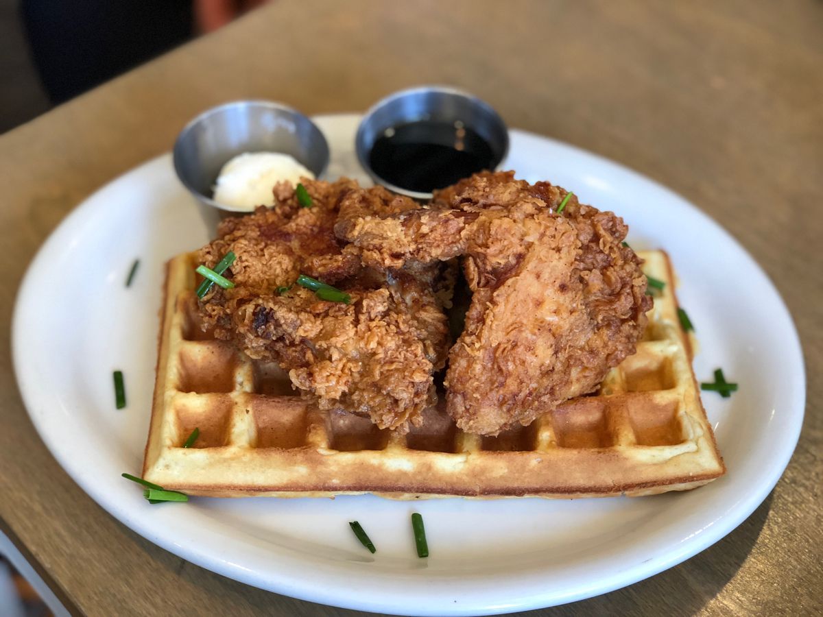 A plate of waffles with fried chicken wings on top. There’s separate dishes of maple syrup and butter served on the side. 