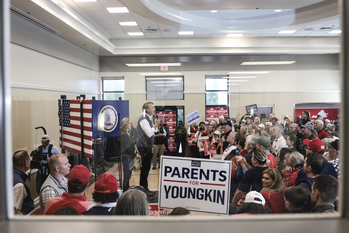 Virginia Governor-elect Glenn Youngkin speaks at an indoor rally, shown behind a glass window. A sign reading “Parents for Youngkin” sits in the foreground by the window.