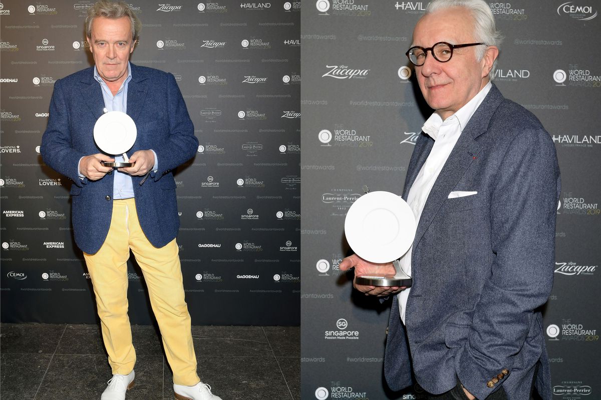 Two white men with white hair, both named Alain, hold a plate-shaped trophy in front of a gray step-and-repeat.