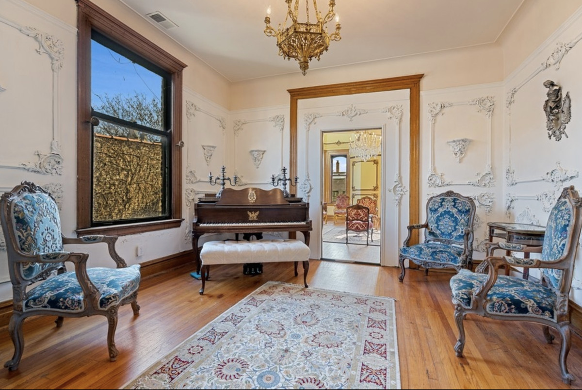 A formal living room with detailed picture molding plaster work, a gold chandelier, blue upholstered chairs, classical decor, and a piano with candelabras on top.