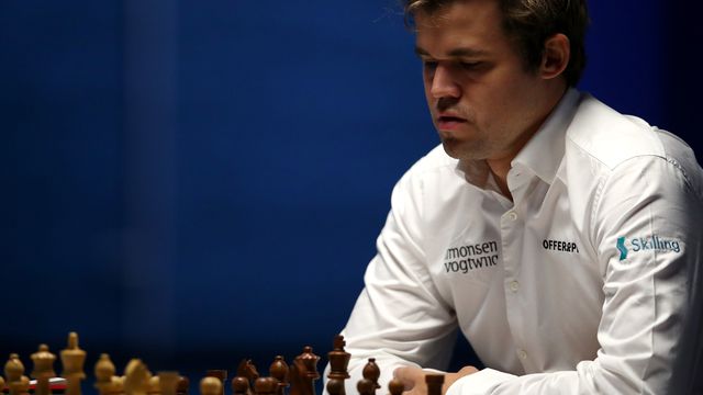 A light-skinned man in a white button down is sitting, looking intently at a chess board populated with pieces. He is contemplating his next move.