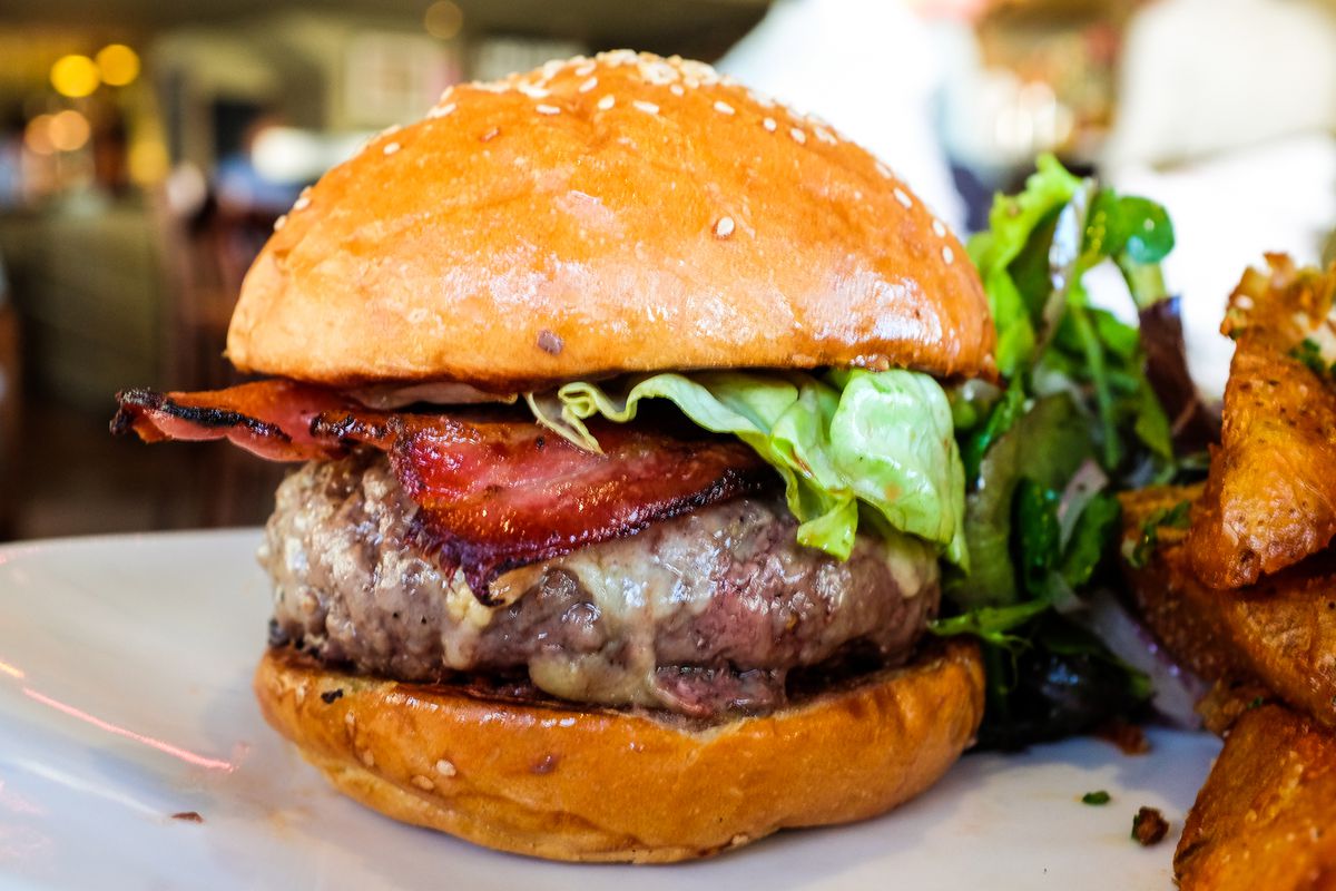 Closeup on a burger featuring a thick patty, bacon, lettuce, and cheese, with thick fries on the edge of the frame