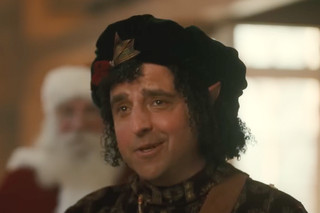 Bernard, o elfo sorrindo para a câmera; Ele é um homem de meia idade com cabelos escuros encaracolados e olhos castanhos quentes, vestindo uma boina verde escura festiva