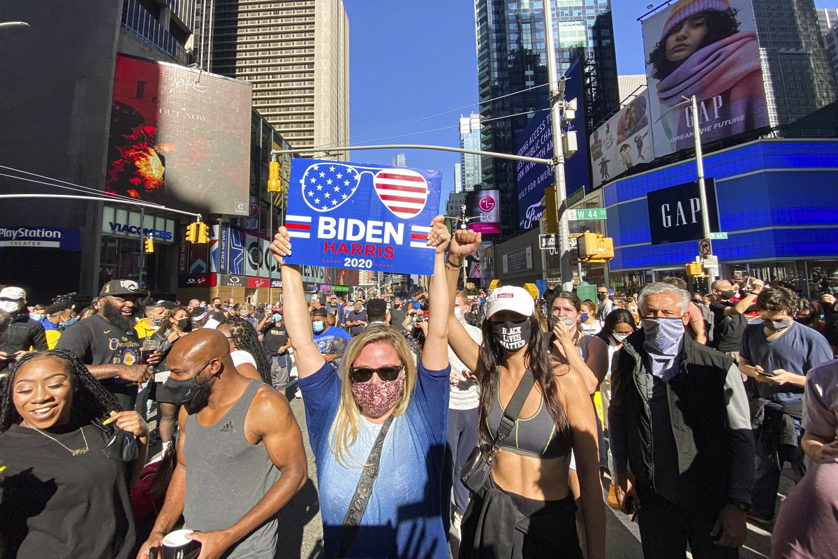 The neon billboards glow in the sunshine above the packed square — all sorts of people, most in masks, celebrate together; the front of the photo has a Black couple, the woman in black, and the man in grey, standing next to a white woman in sunglasses, holding a sign above her head that says ‘Biden Harris 2020,’ that features Biden’s trademark aviators. Beside her is a woman in a mask that reads ‘Black Lives Matter,’ her fist raised to the sky.
