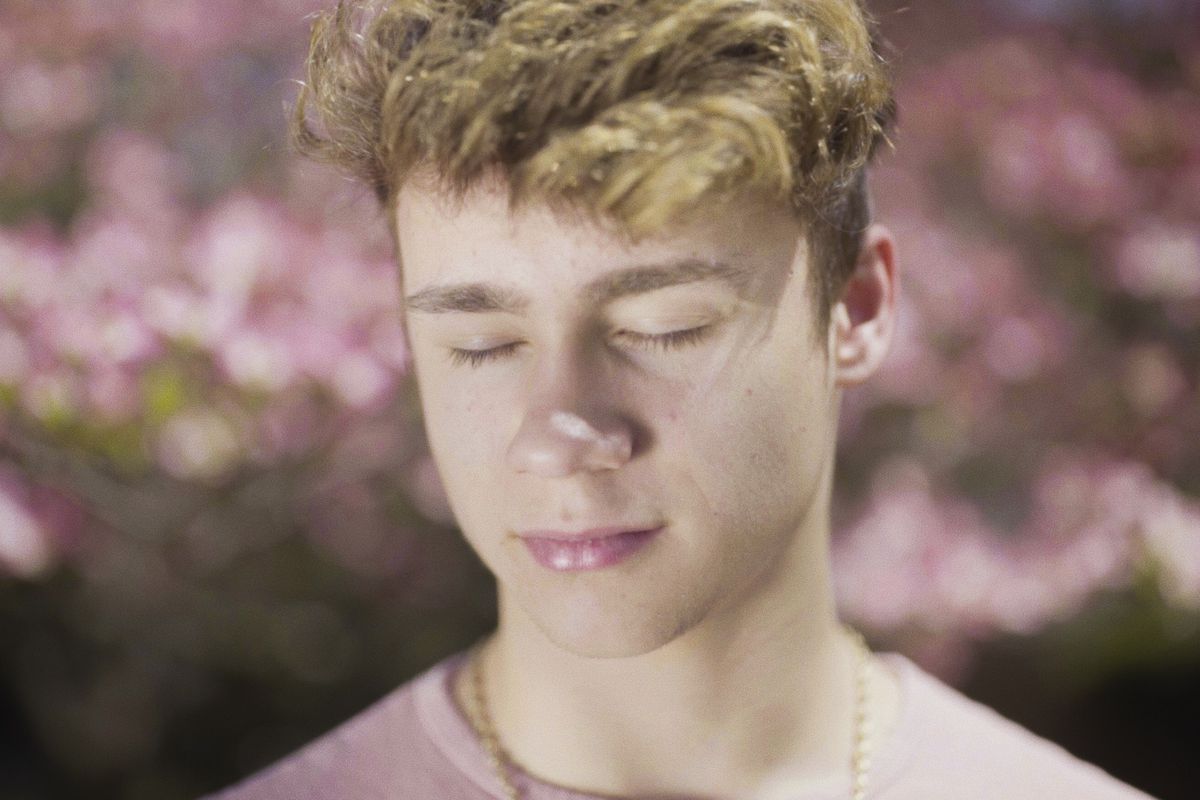 Austyn Tester, the young protagonist of the documentary “Jawline,” stands in front of a pink backdrop with his eyes closed.