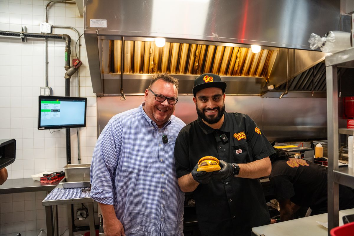 Chris Shepherd smiles alongside Burger Bodega founder Abbas Dhanani, who holds a burger.
