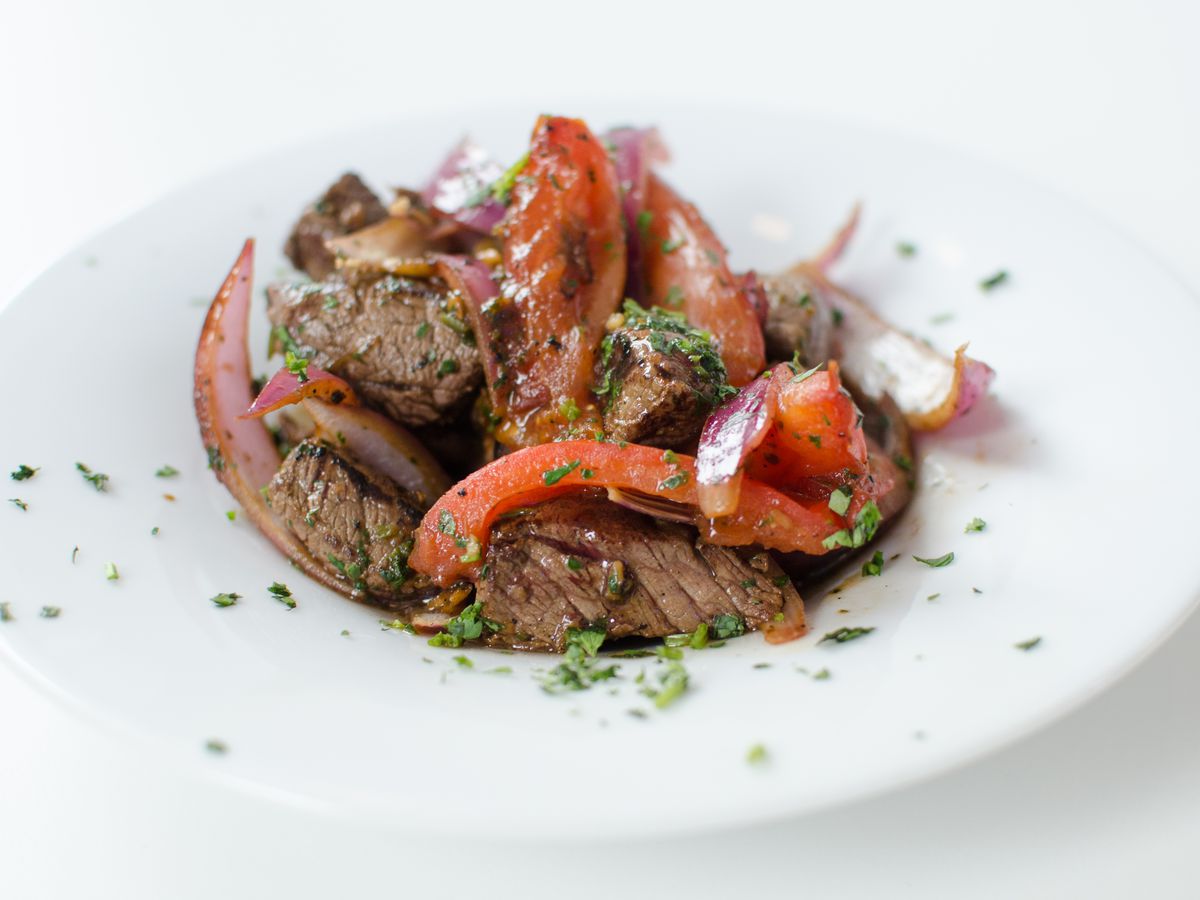 A white bowl on a white table is full of chunks of cooked beef, tossed with slices of red pepper and tomatoes. The dish is topped with green herbs.