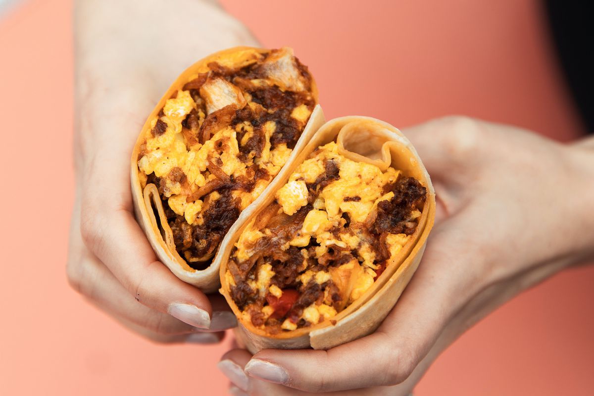 Two hands hold a breakfast burrito, split open, over an orange table.