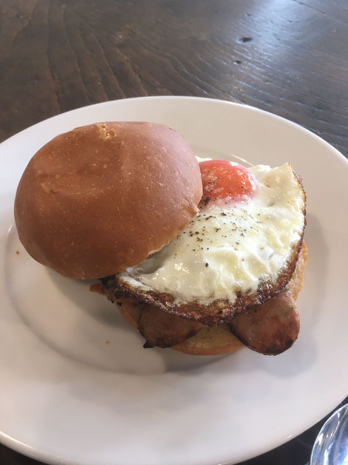 A bread roll filled with fried egg and sausages on a white plate, on a wooden table.