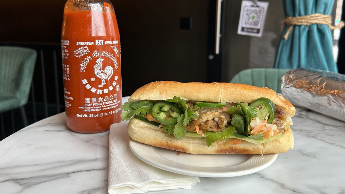 A banh mi and a bottle of Sriracha hot sauce sit on a marble table at Coconana.