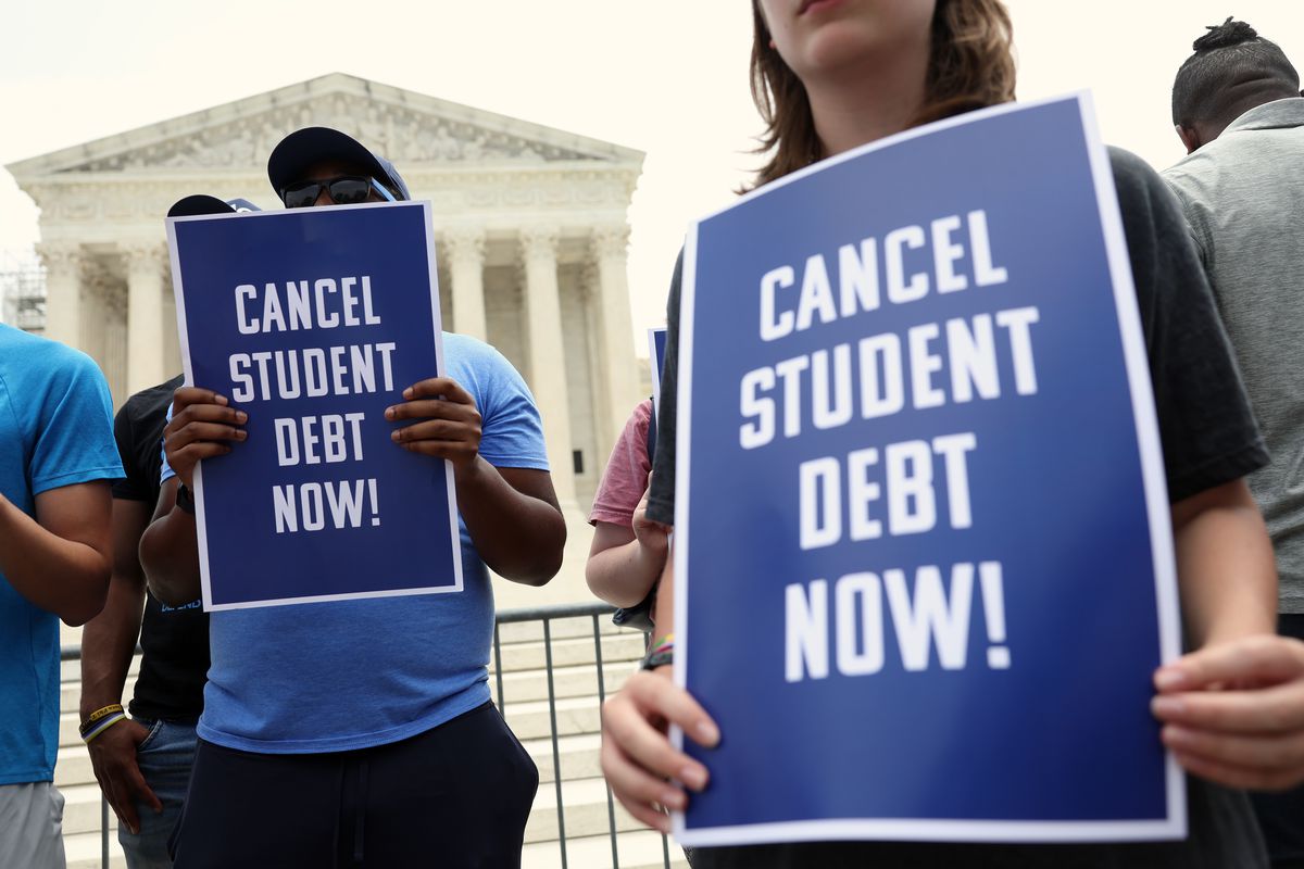 Two activists, holding blue signs bordered in white with white text reading “Cancel student debt now!,” stand in front of the US Supreme Court building.