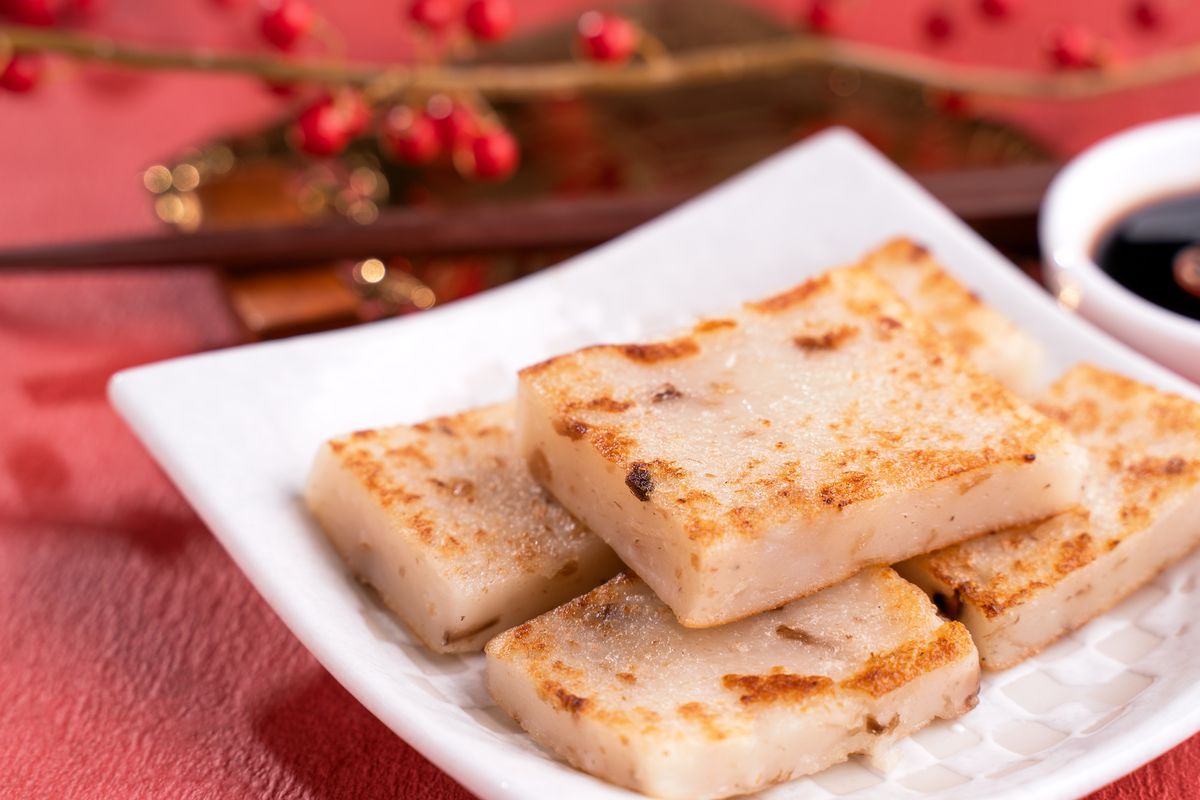 Several pieces of luóbo gāo (turnip cake) have been arranged on a square white dish that sits atop a red tablecloth.