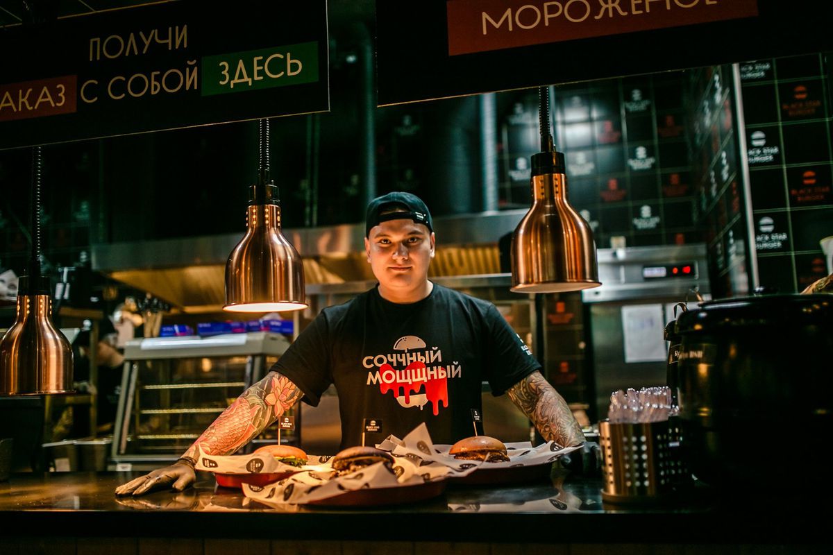 A dark photo of a man in a Russian shirt looking out over a tray of burgers inside a restaurant.