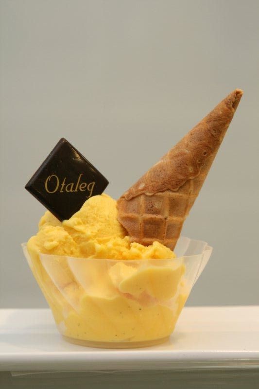 A plastic dish filled with bright yellow gelato with a cone sticking out to one side, and a chocolate square branded with “Otaleg” to the other