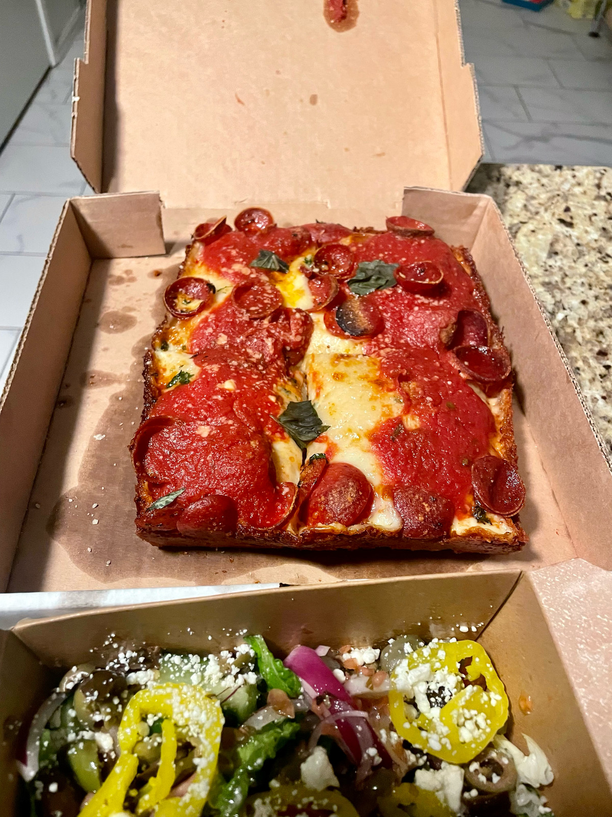 In the back, a pepperoni deep dish pizza sits in an open delivery box. In the foreground is a Greek salad with vibrant green banana peppers.
