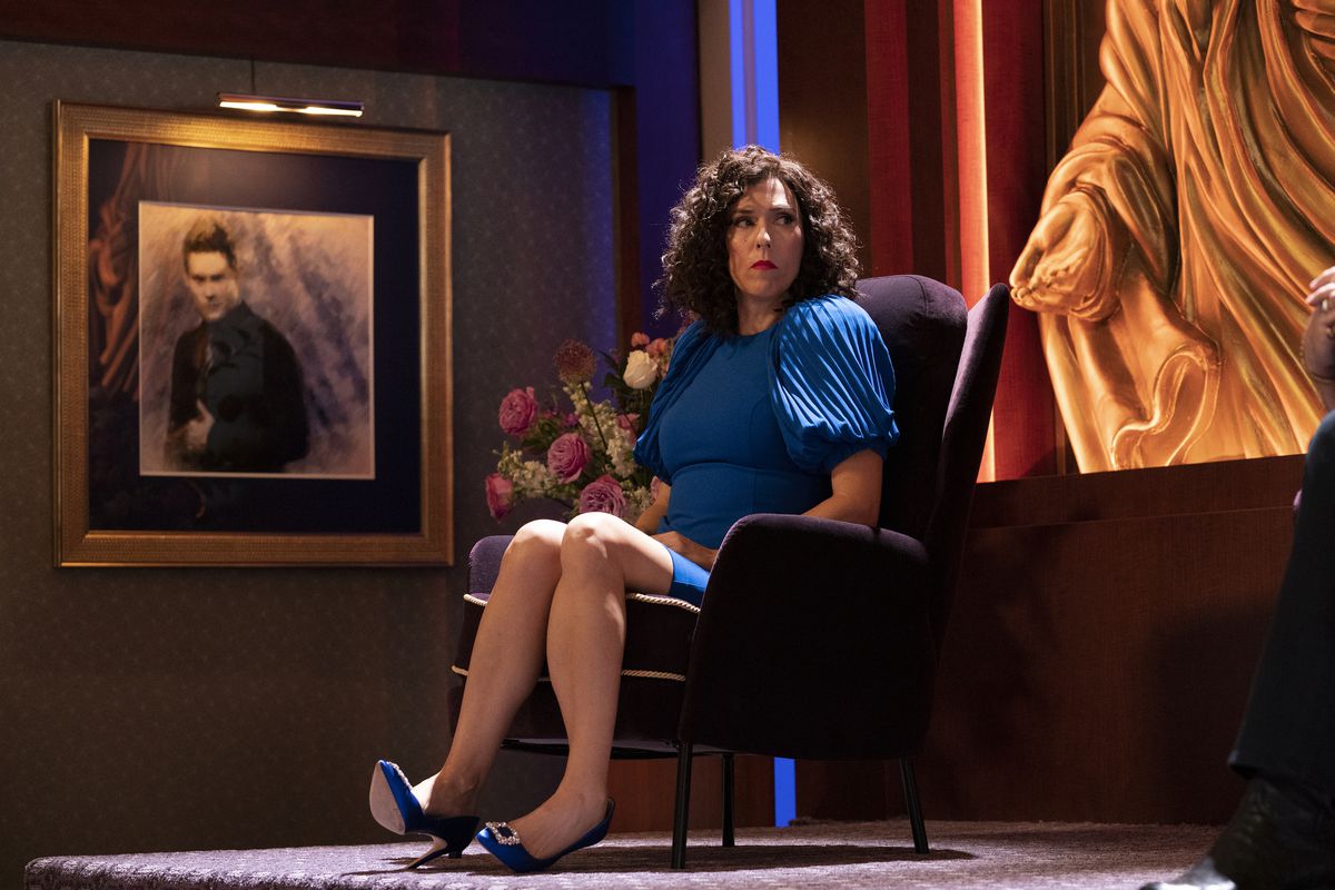 Judi Patterson, wearing a blue dress with blue heels, sits on a purple chair on a raised platform in The Righteous Gemstones. Behind her is a giant statue of herself, while to the right is a sketched framed drawing of Kelvin Gemstone (Adam Devine).