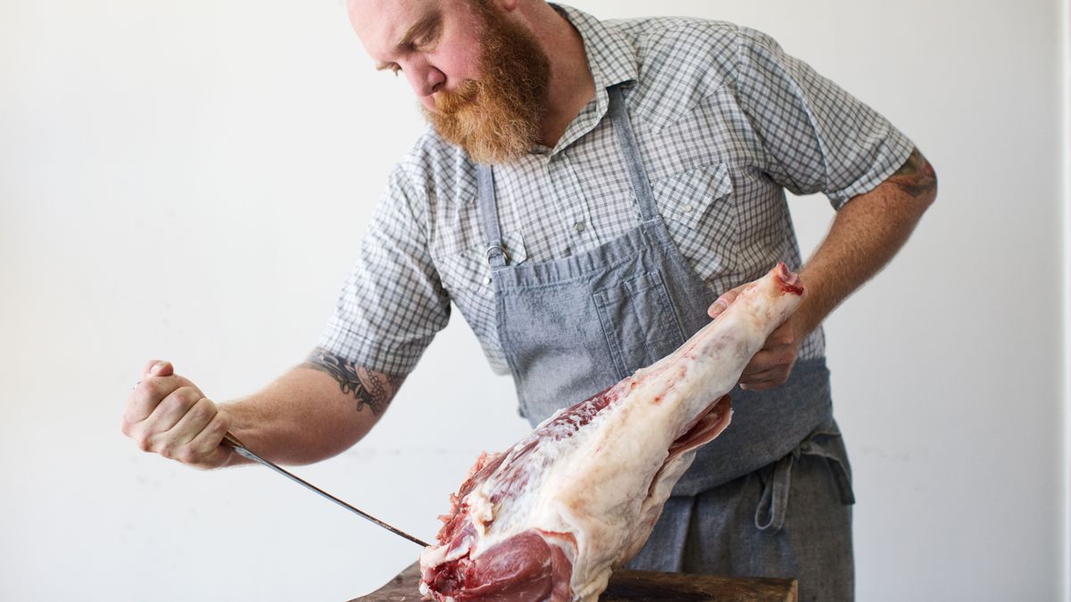 A bearded man in a stripped light-colored shirt and a light blue apron holding a knife to a boar leg