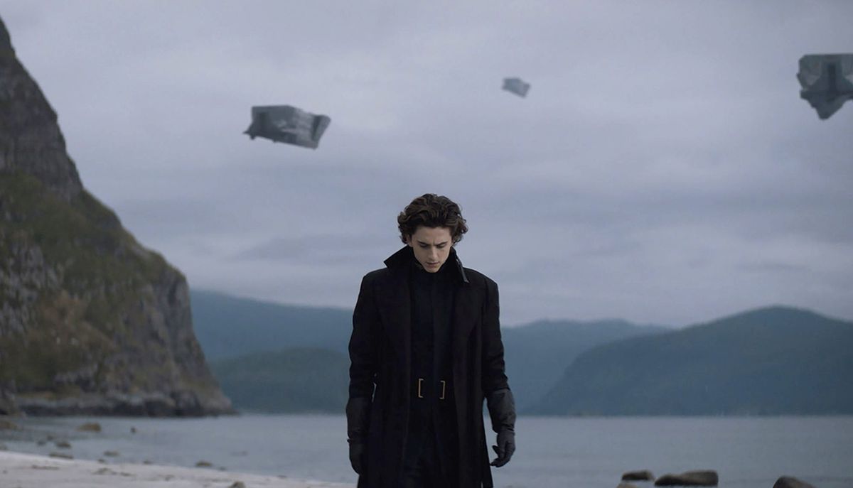 A young man in all-black clothing walks on a beach, gray skies and flying ships in the air above him.