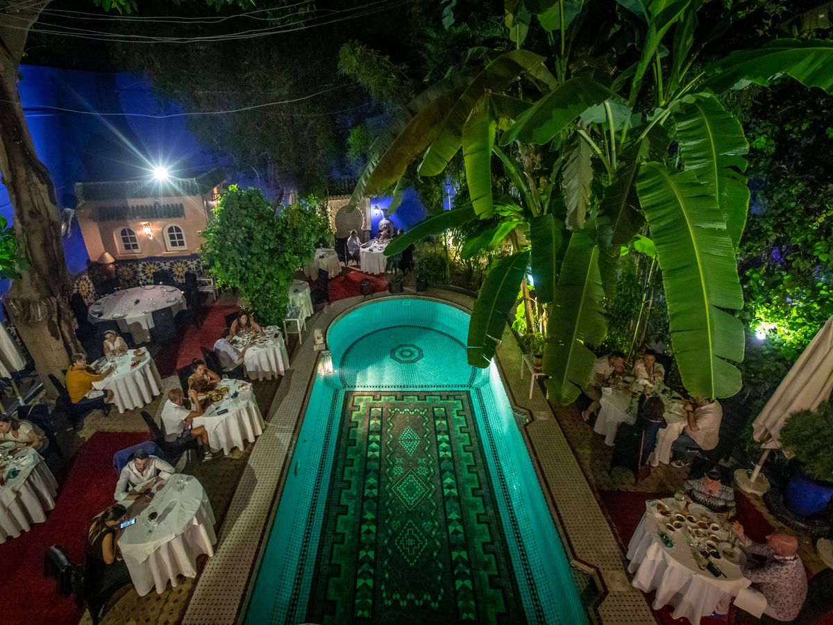 From a high perch, a large pool with a decorative floor in the middle of an interior courtyard/dining room where guests are seated at tables with white tablecloths, large palm trees reach toward the camera, and small structures imitate exterior buildings