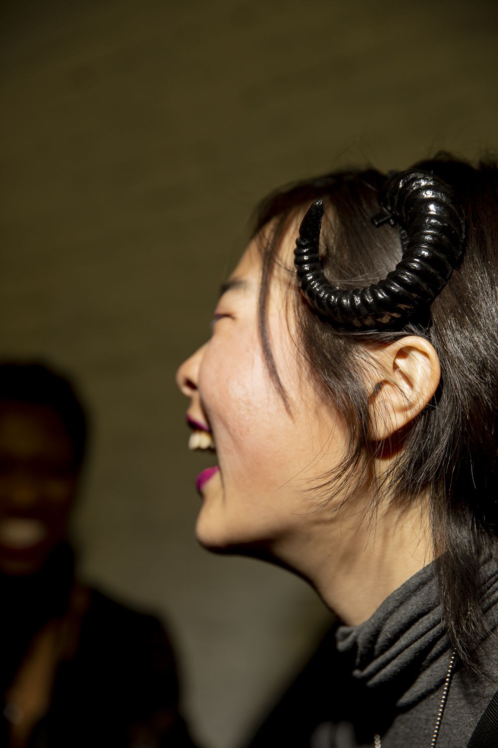 Close-up of a person laughing in profile, a black plastic ram’s horn curling off of a headband, just over her left ear.