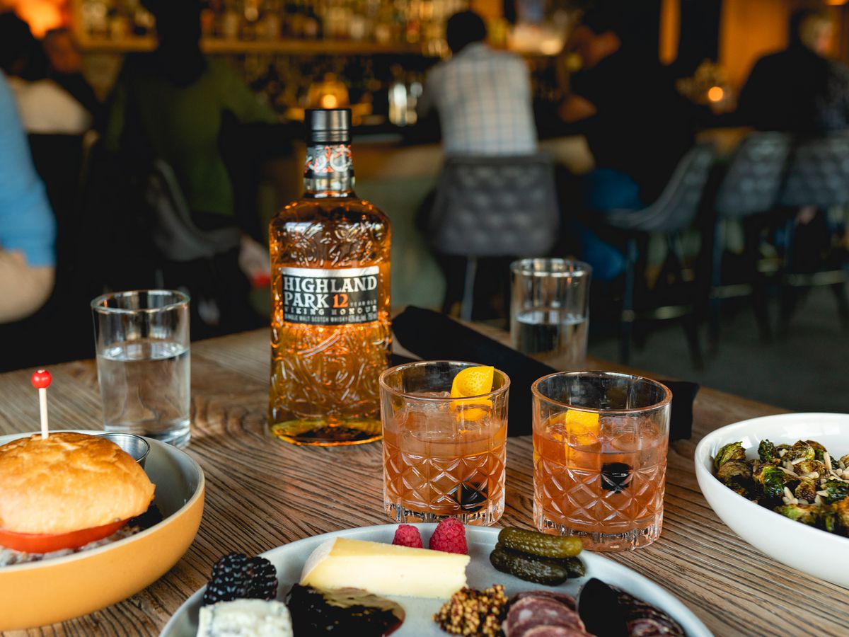 A table with two cocktails, a bottle of Highland Park whisky, and plates of charcuterie, salad, and a sandwich.