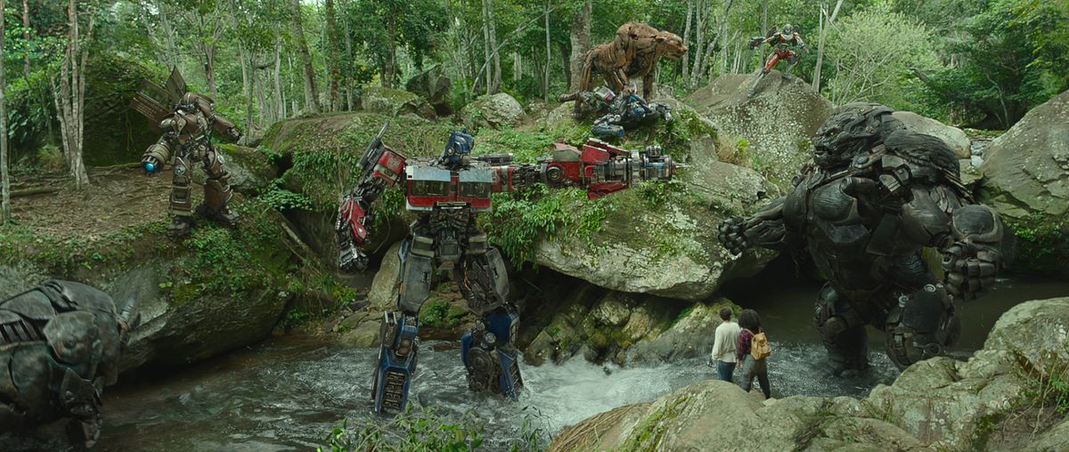 Optimus Prime (a big red-and-blue humanoid robot) points a gun-arm in the face of Optimus Primal (a big black gorilla-shaped robot) while standing in a stream outside a forest cave. Members of their robot factions, in humanoid or animal form (Rhinox, Wheeljack, Mirage, Cheetor, Arcee) array around them, with Anthony Ramos and Dominique Fishback as tiny human figures dwarfed by the giant robots, in Transformers: Rise of the Beasts