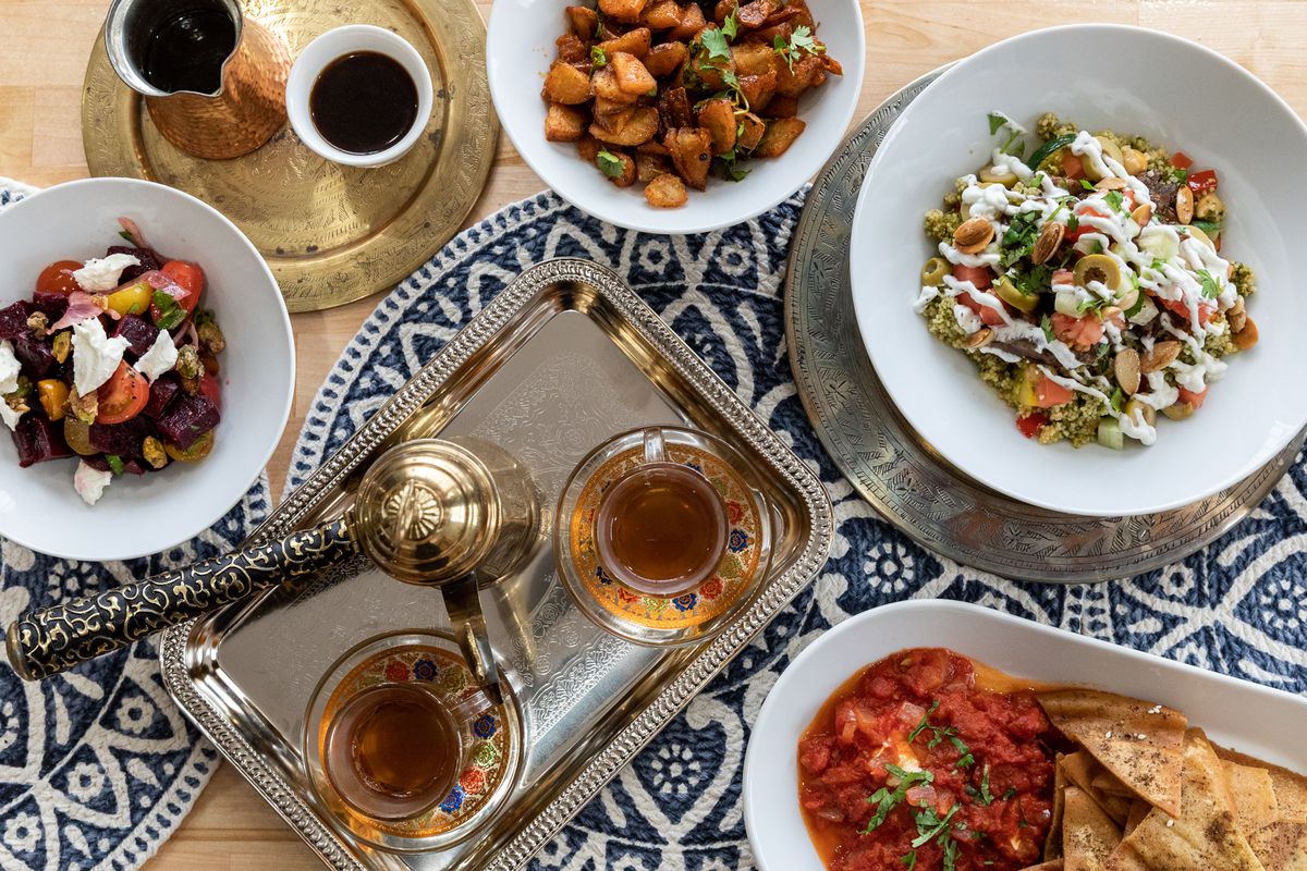 A Moroccan tea set, a red dip with pita chips, a lamb tagine, harissa potatoes, coffee, and more on a wooden table with blue and white-patterned woven center pieces.
