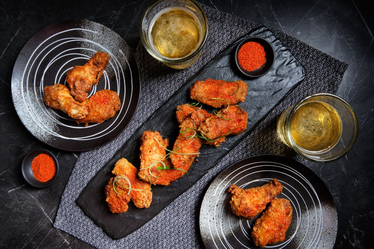 Togarashi hot honey fried chicken wings are served on a rough-hewn black slab, surrounded by small plates of wings and two glasses of beer.