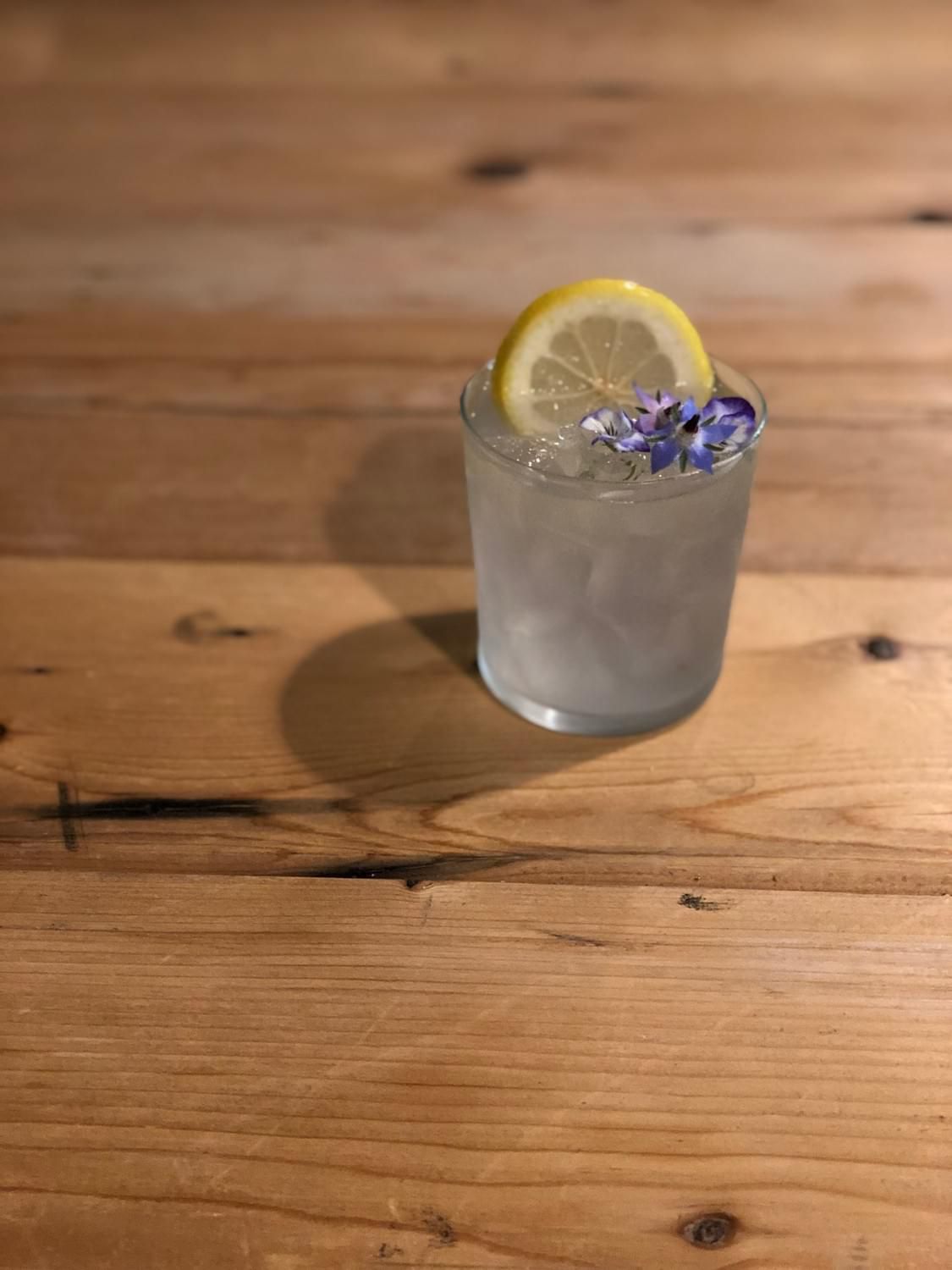 A clear highball glass filled with pale, clear liquid and adorned with a purple flower and lemon slice sits atop a light wood surface