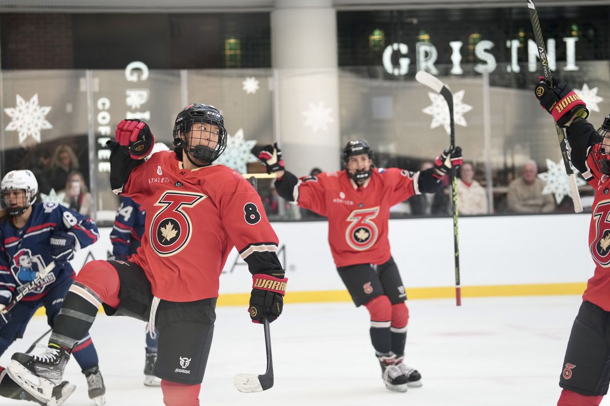 Toronto Six player Leah Lum celebrates a goal in front of two of her teammates and two Riveters player. She has her right arm in the air and her right leg raised as she is about to skate past the bench