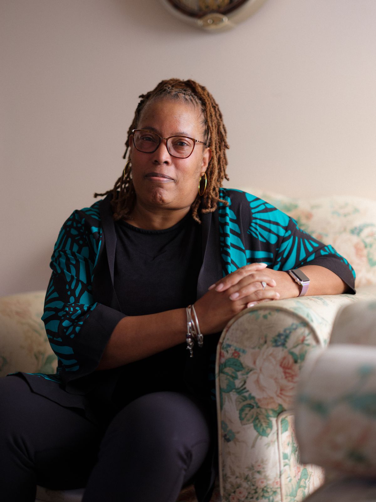 A Black woman sits on a cream floral-patterned couch, wearing a teal and black sweater and glasses.