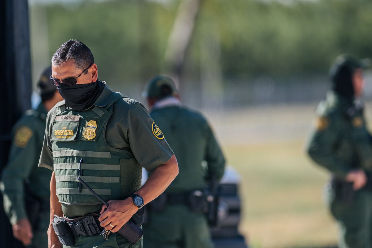 A boarder patrol agent stands with sunglasses and a mask on his face and armor on his body.