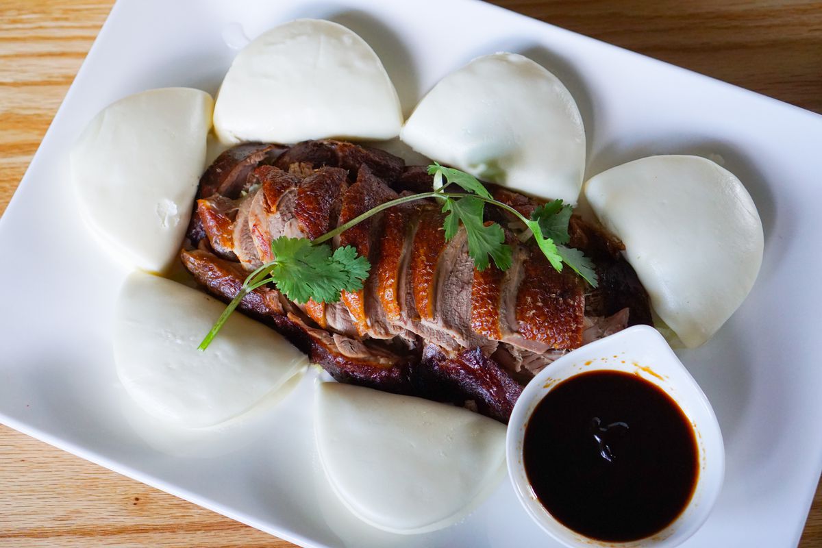 A plate filled with sliced smoked tea duck  topped with cilantro and steamed buns, with a side of dipping sauce.