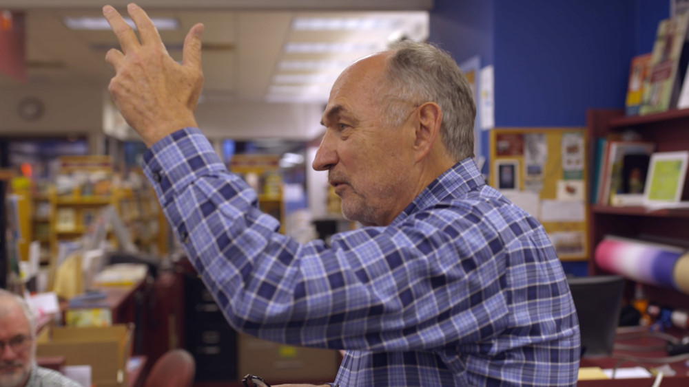 An old man in a checkered blue shirt raises his hand and speaks in a bookstore.