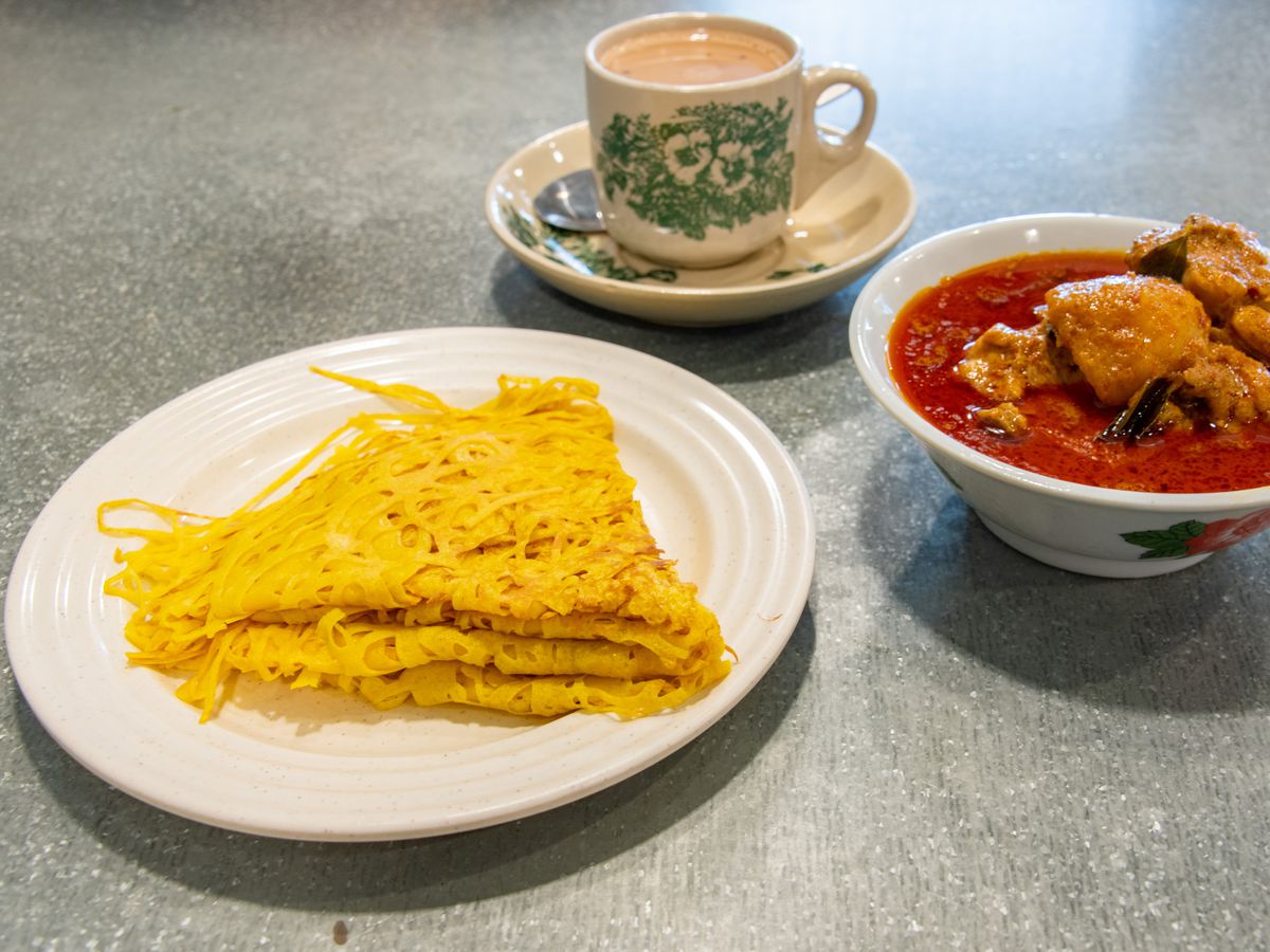 A plate of roti jala, beside a cup of tea in traditional kopitiam mug and a stew