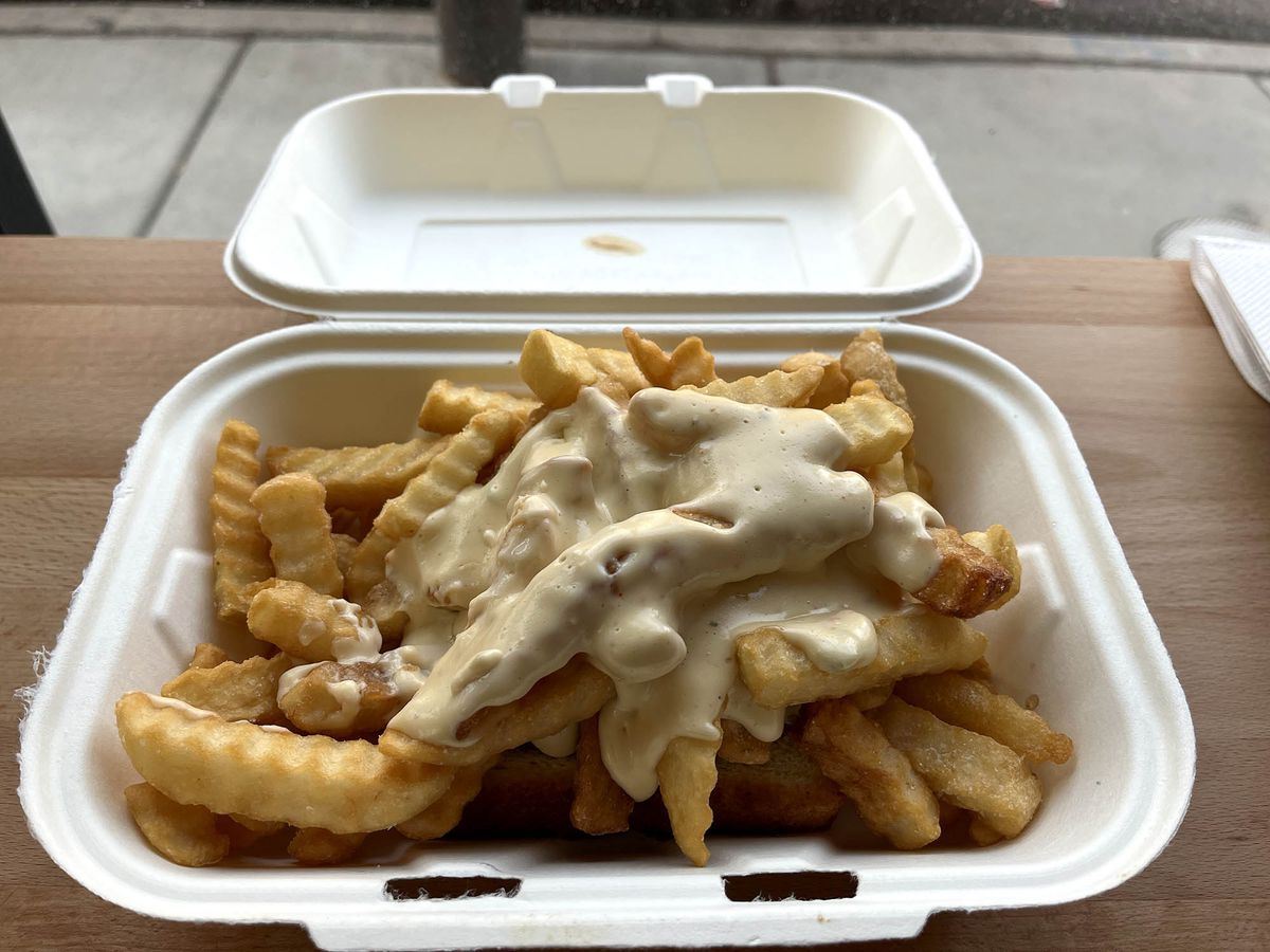 A cardboard to-go box filled with a heap of fries on a slice of toast, covered in pale yellow cheese sauce.