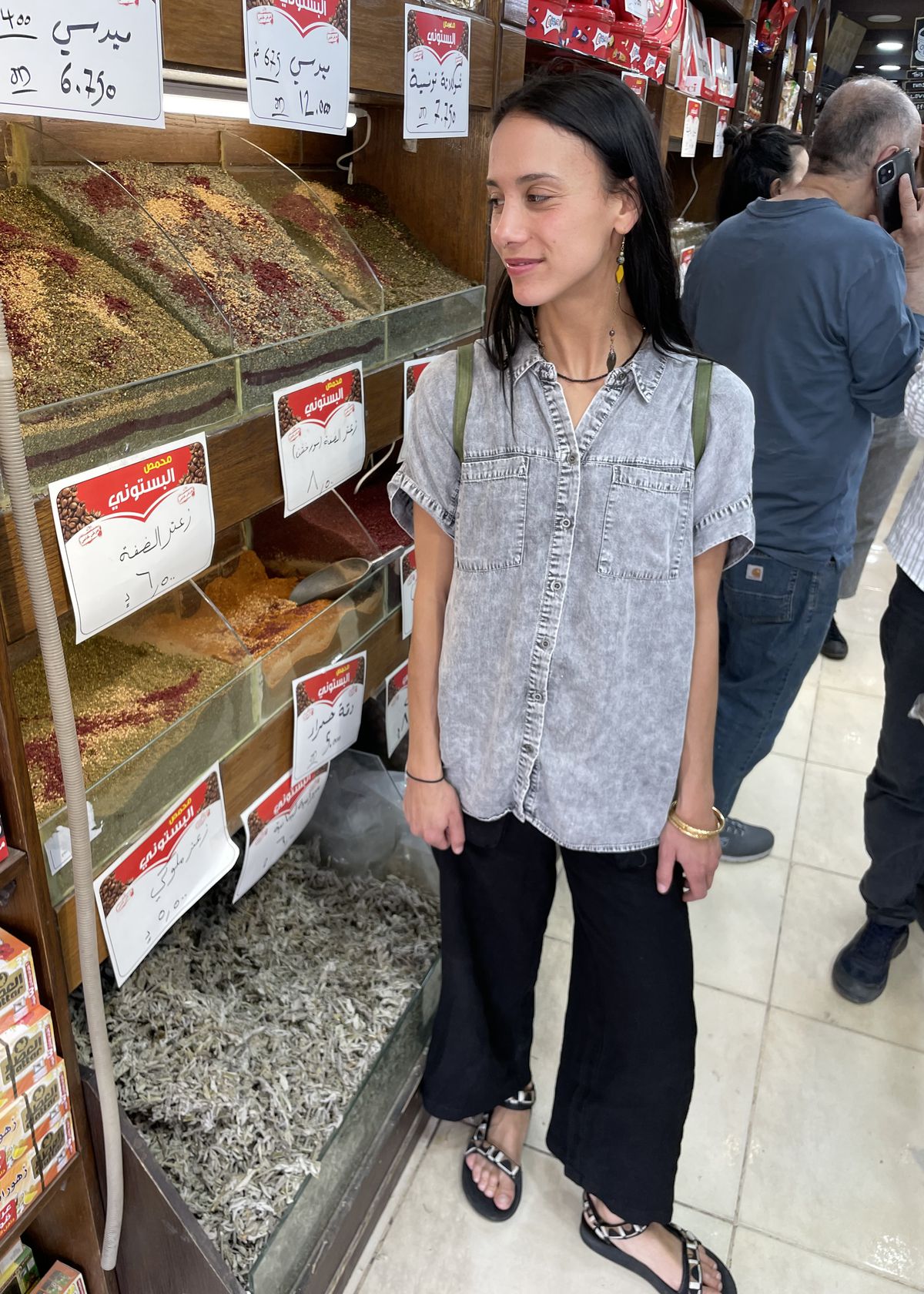 Lyla Abukhodair, wearing black pants and a grey button-down shirt, standing next to shelves of spices in a shop.