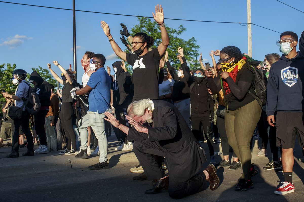 A diverse group of protesters, all in masks, hold up both hands; and older gentleman with white hair raises his hands as he takes a knee.