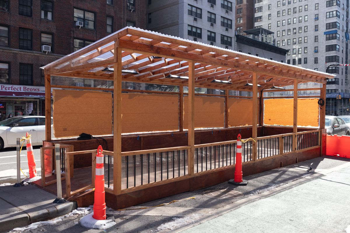 A plywood structure on the street with orange traffic cones placed around it.
