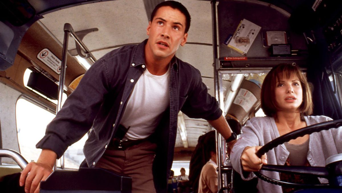 A man with low-cropped black hair (Keanu Reeves) in a blue denim dress shirt and white undershirt stands beside a woman (Sandra Bullock) driving a bus.