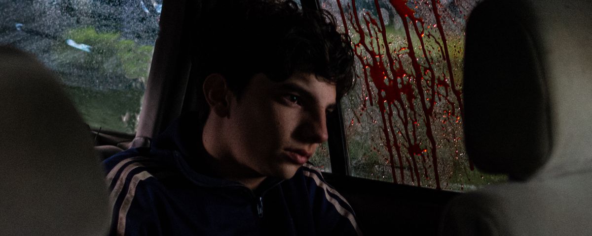 A teenager in a track suit with a fixed, blank expression on his face sits in the back seat of a car at night, next to a window thickly smeared with dripping red blood in When Evil Lurks