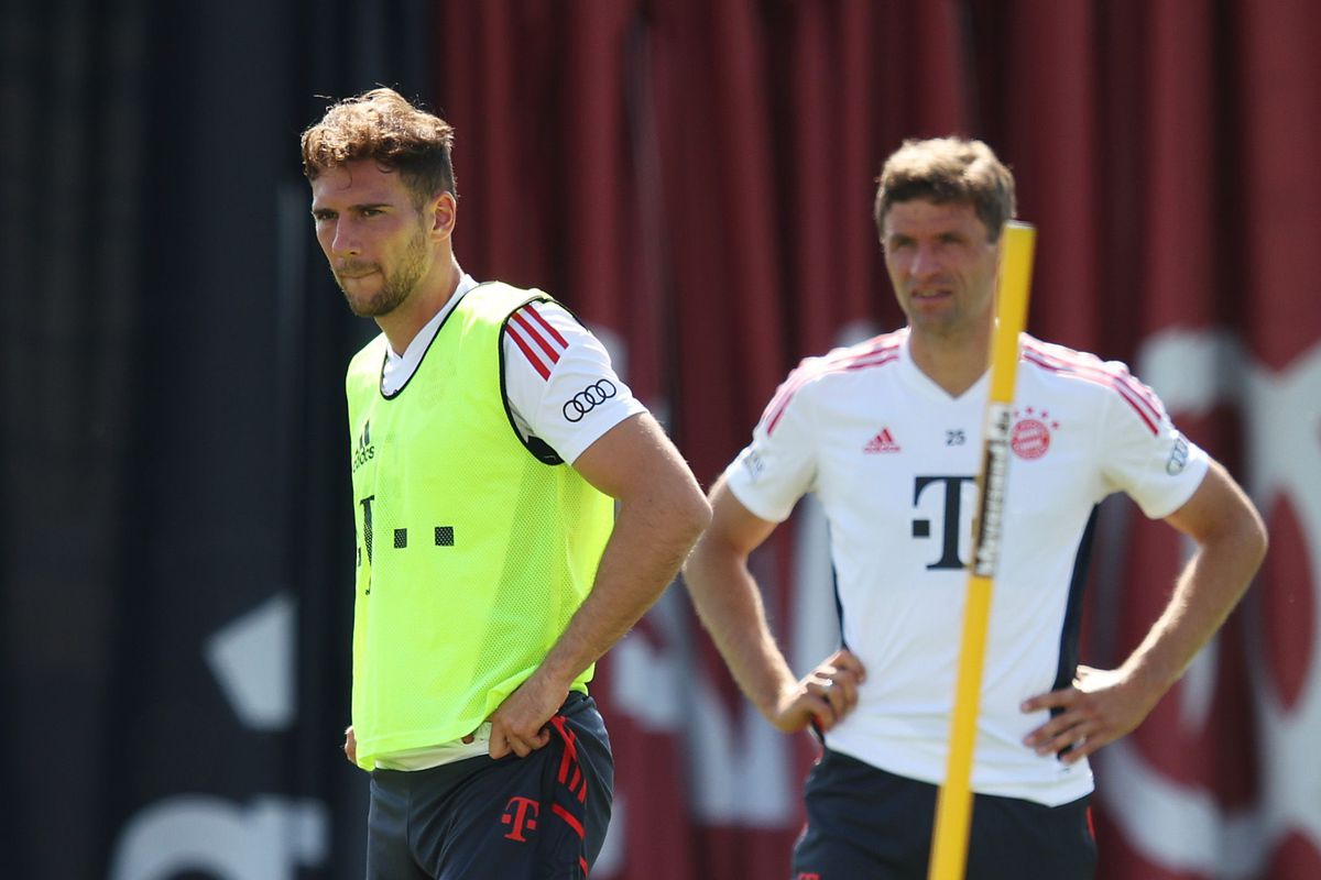 Leon Goretzka, in a yellow practice bib, stands on the field during training as Thomas Müller looks on in the background.