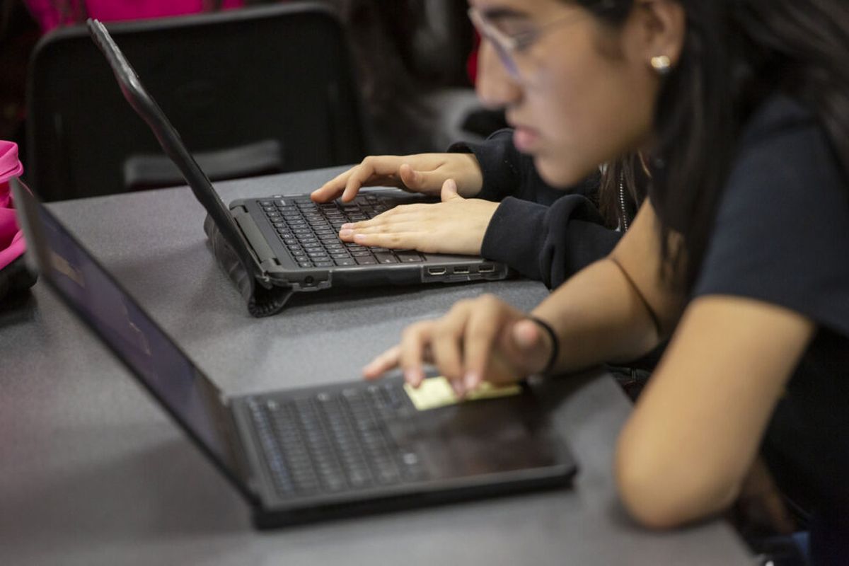 A high school student at DCIS Montbello leans toward a laptop on a table and looks intently into the screen. The hands of another student sitting to the right are seen on a laptop. The students were working on an assignment in class in May 2019.